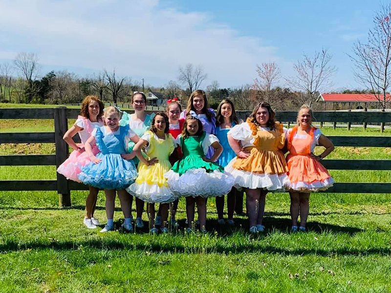 Group photo of lincoln county cloggers dressed in colorful dresses standing in the grass against a wooden fence
